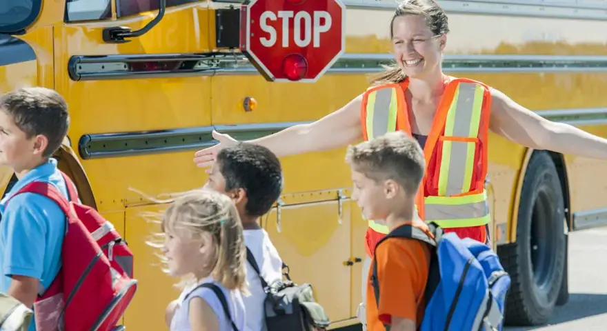 Des enfants montent dans un bus scolaire jaune, guidés par une adulte portant un gilet de sécurité orange.