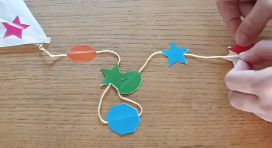 Fabrication d'un cerf-volant étoile : fils, formes colorées (étoiles, ovales, octogone) et mains.
