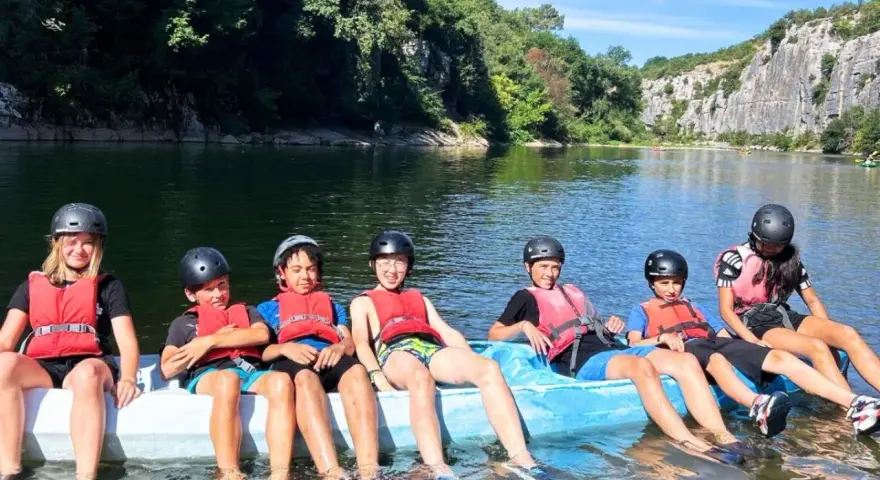 Des enfants en gilets de sauvetage et casques sur un raft bleu sur une rivière, entourés de verdure et de falaises.