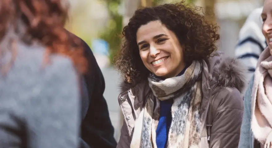 Une femme souriante aux cheveux bouclés porte une écharpe et une veste à capuche en fourrure.