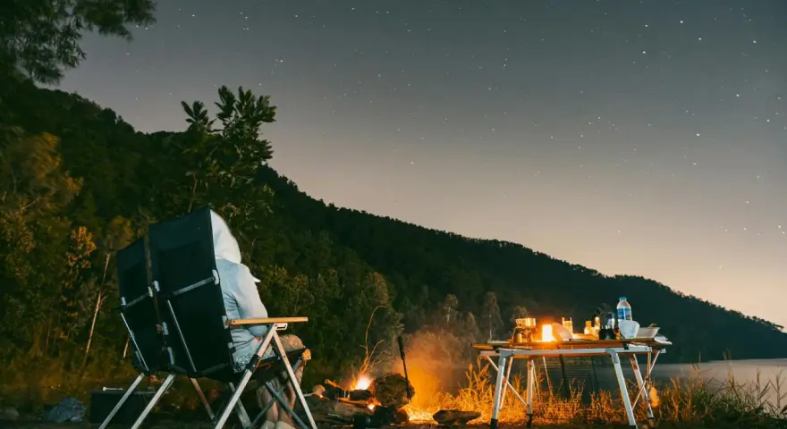 Personne assise près d'un feu de camp sous un ciel étoilé, avec une table de camping et un lac.