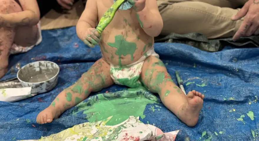 Bébé couvert de peinture verte, tenant un pinceau, assis sur une bâche bleue.