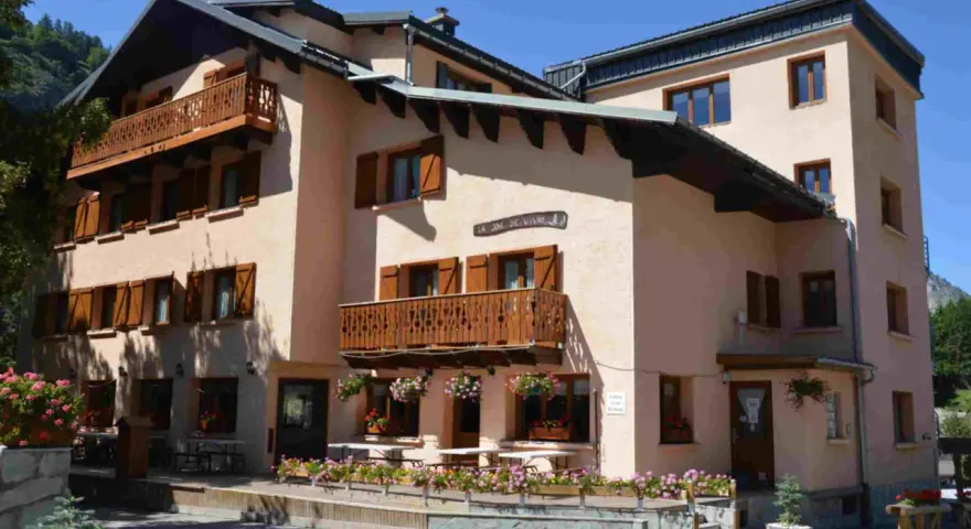 Bâtiment de style alpin avec balcons en bois et volets marrons, fleuri. Une enseigne "La Joie de Vivre" suggère une ambiance de colonie en Haute-Savoie.