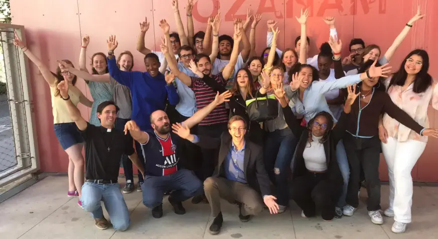 Un groupe de jeunes gens, dont un prêtre, pose joyeusement devant un mur rose avec le texte "MAISON OZANAM".
