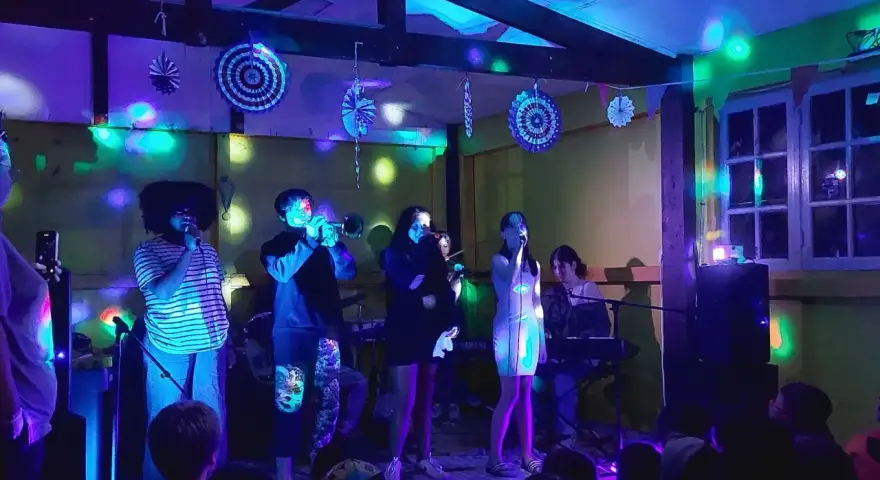 Scène de concert animée avec des lumières colorées. Un groupe joue, avec des chanteurs, un trompettiste et un claviériste. Le public, dont des enfants, filme le spectacle. Ces vacances musicales sans frontières sont une fête pour les sens.
