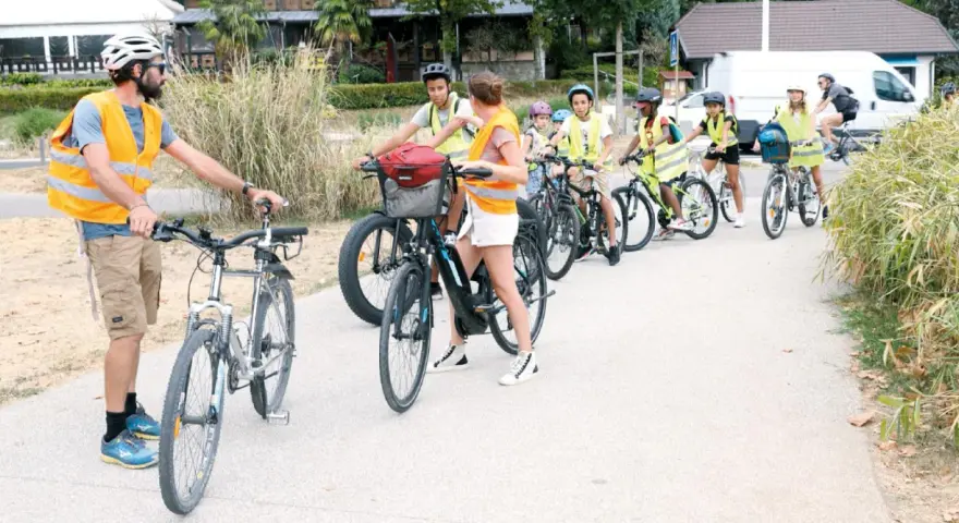 Un groupe d'enfants à vélo, encadrés par des adultes en gilets de sécurité, suit un parcours. La scène illustre la réglementation du déplacement à pied et à vélo à l'ACM.