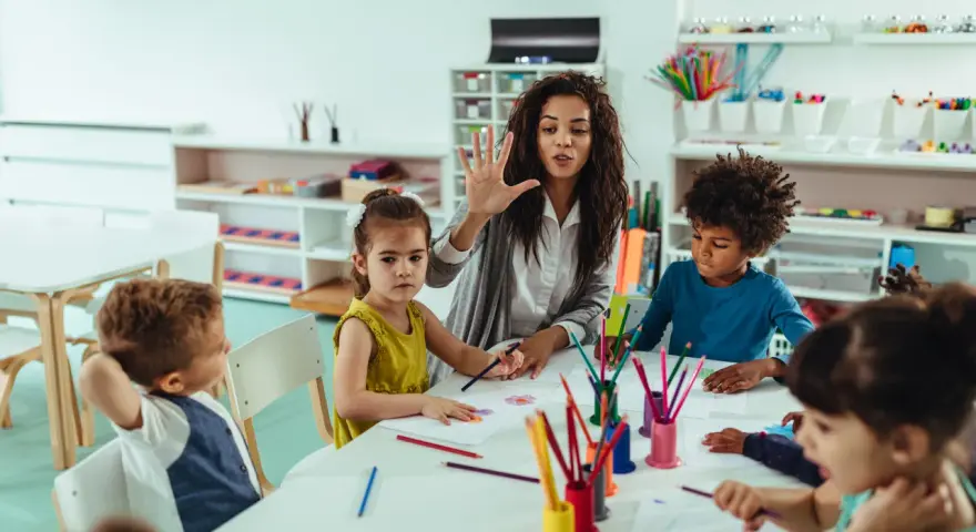 Une animatrice explique une activité créative à des enfants autour d'une table. Son rôle est d'éveiller leur curiosité.