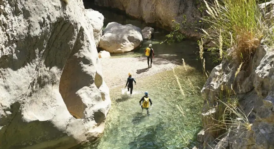 Trois personnes en combinaison font du canyoning dans une rivière aux eaux claires, entourées de falaises rocheuses.