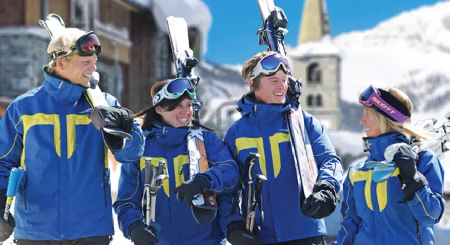 Quatre jeunes skieurs souriants en tenue bleue et jaune, portant des skis et des bâtons, dans une station de ski enneigée.