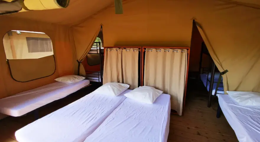 Intérieur d'une chambre de colonie de vacances avec plusieurs lits simples, draps blancs et oreillers. Des rideaux séparent les espaces.