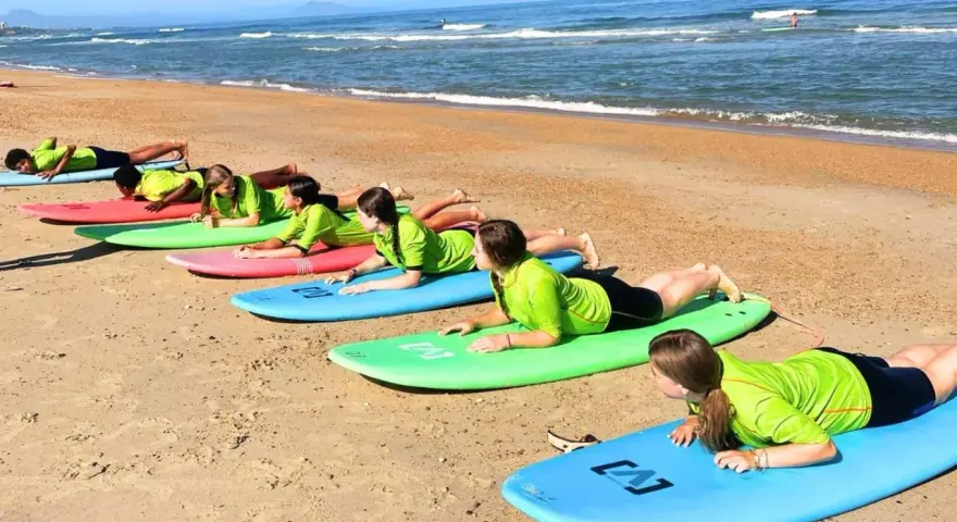 Des adolescents en stage sportif sur la plage, allongés sur des planches de surf, apprenant les bases avant de se jeter à l'eau.