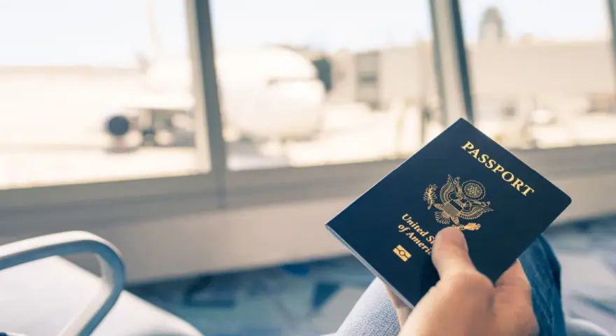 Main tenant une carte d'identité américaine devant une fenêtre d'aéroport avec un avion en arrière-plan.