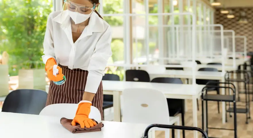 Employée portant masque et visière nettoie une table, respectant la réglementation hygiène cantine scolaire. Des séparateurs sont installés.