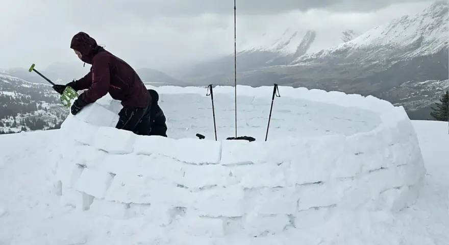 Person construisant un igloo en blocs de neige, avec des bâtons de ski plantés.