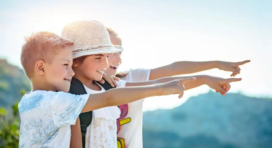Trois enfants souriants, portant des sacs à dos, pointent du doigt vers la droite, sous un ciel ensoleillé.