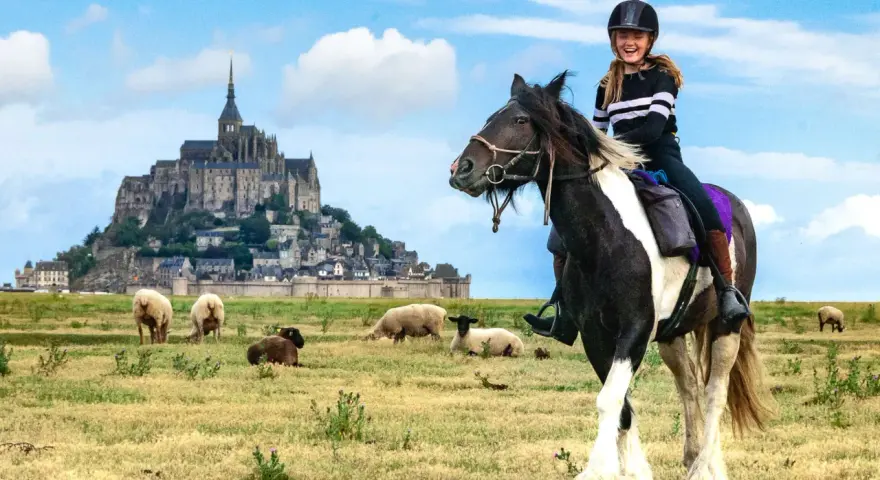 Une jeune fille rit à cheval devant le Mont-Saint-Michel, avec des moutons dans un pré.