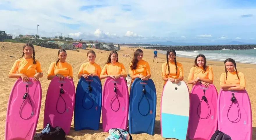 Huit jeunes filles en t-shirts orange, prêtes pour leur colonie à la mer, posent avec leurs bodyboards sur la plage.