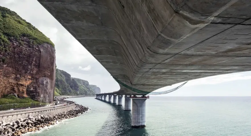 Vue sous un pont en béton, avec la mer, des falaises verdoyantes et une route côtière.