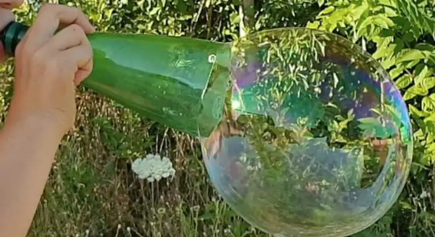 Main d'enfant soufflant dans une bouteille verte pour créer une bulle de savon géante irisée.
