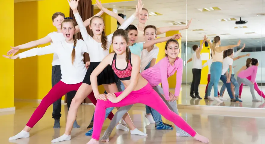 Une joyeuse colonie de danse, composée d'enfants et d'un professeur, s'exerce avec enthousiasme dans un studio lumineux aux murs jaunes.