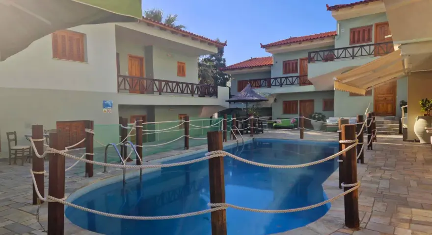 Piscine bleue entourée de cordes et de poteaux en bois, avec des bâtiments de style colonie en Grèce en arrière-plan.