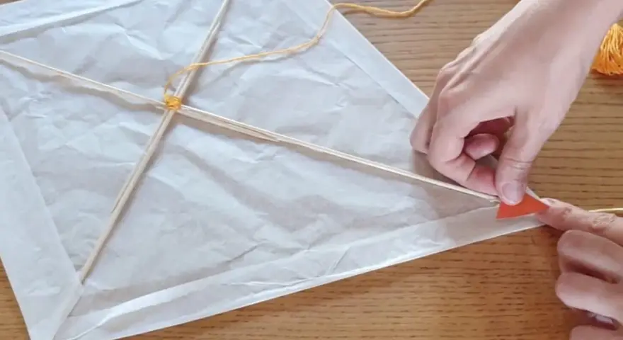 Mains fabriquant un cerf-volant simple en papier blanc et bâtons de bois, avec fil jaune.