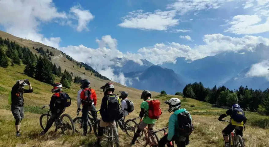 Groupe d'ados en colonie VTT descente, prêts à dévaler les pentes montagneuses sous un ciel bleu éclatant.