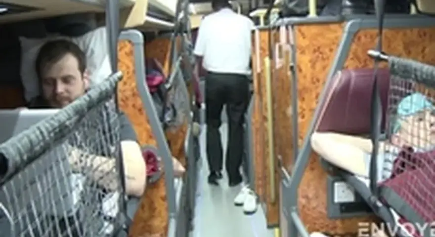 Intérieur d'un bus avec des couchettes. Un homme en blanc marche dans l'allée, tandis que des passagers se reposent dans leurs couchettes.