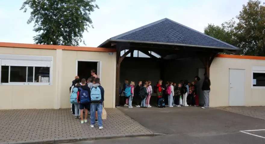 Des enfants font la queue devant un bâtiment scolaire, certains avec des sacs à dos.