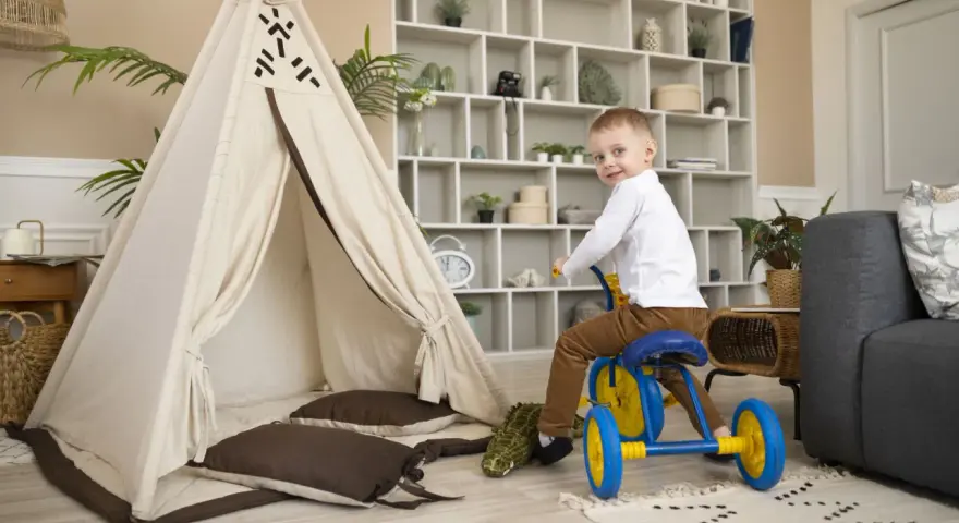 Un petit garçon sur un tricycle bleu et jaune s'amuse dans un espace de jeux, près d'une tente tipi et d'un canapé. Ce centre de vacances et de loisirs est idéal pour les enfants.