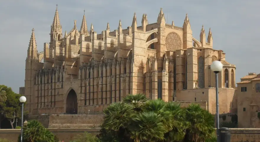 La cathédrale de Palma, majestueuse, se dresse sous un ciel d'hiver. Des palmiers verdoyants bordent le chemin, offrant un contraste saisissant avec la pierre dorée du monument.