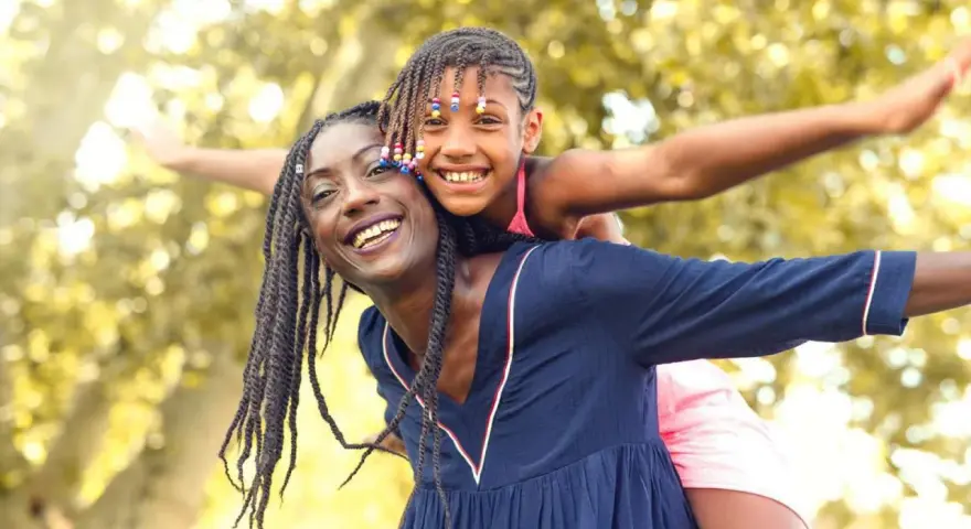 Une femme porte une enfant sur son dos, toutes deux souriantes, avec des arbres en arrière-plan.