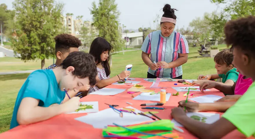 Des enfants créent des œuvres d'art en plein air autour d'une table recouverte d'une nappe orange.