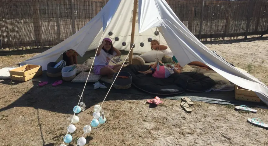 Enfants profitant d'une colonie de vacances camping dans une tente tipi, entourés de coussins et de guirlandes lumineuses.