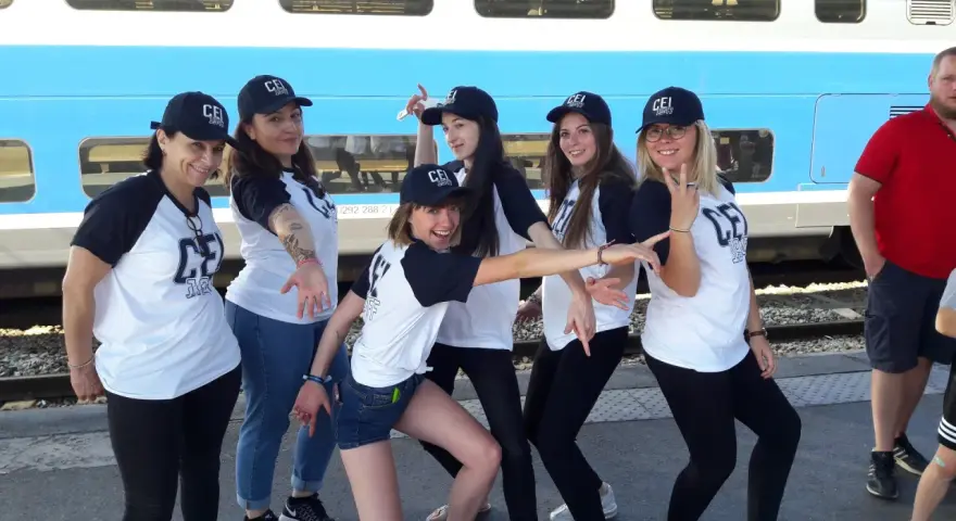 Groupe de jeunes femmes souriantes portant des casquettes et t-shirts "CEI 1947" devant un train.