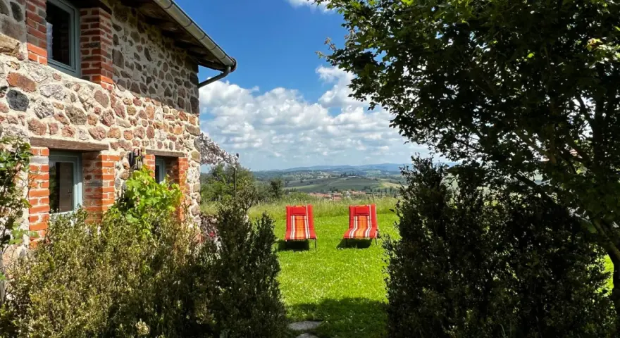 Maison en pierre avec vue panoramique sur les collines, deux chaises longues rouges invitent à la détente. Parfait pour découvrir les destinations Rhône-Alpes.