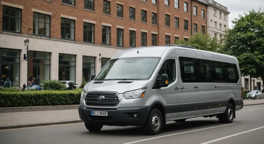 Un homme est au volant, prêt à conduire un minibus argenté dans une rue bordée d'arbres et de bâtiments.