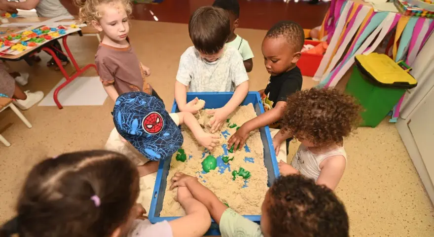 Des enfants s'amusent dans un bac à sable au centre de loisirs. Un petit garçon porte une casquette Spiderman. Ils ont tous environ 2 ans.
