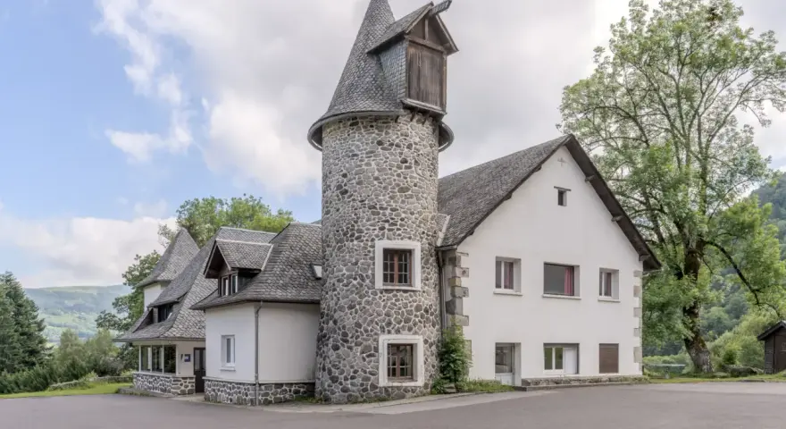 Bâtiment blanc avec une tour ronde en pierre et un toit en ardoise, entouré d'arbres et de verdure.
