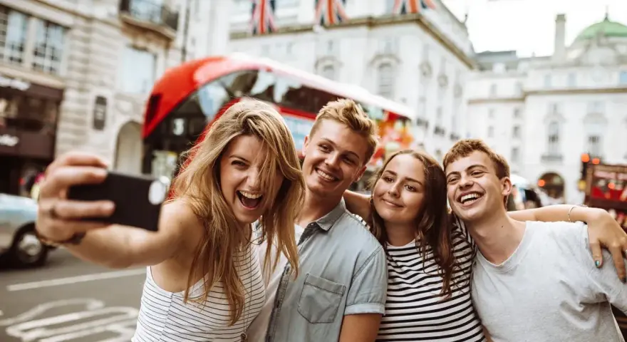 Quatre jeunes souriants prennent un selfie à Londres, avec un bus rouge et des drapeaux britanniques en arrière-plan.