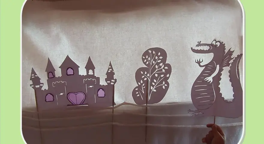 Théâtre d'ombres : château, arbre et dragon en papier, manipulés par une main.