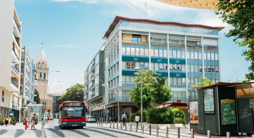 Un bus rouge circule dans une rue bordée d'immeubles et d'un clocher, sous un ciel bleu.