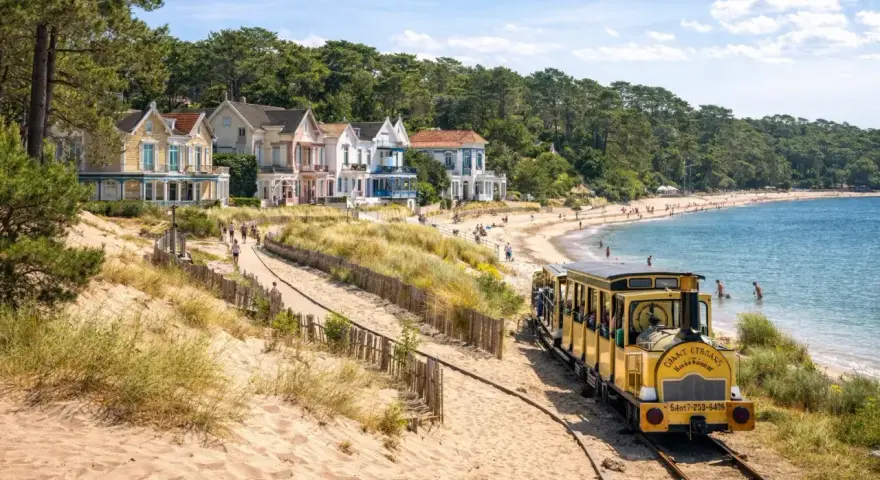 Petit train jaune longeant une plage de sable bordée de maisons colorées et d'une forêt.