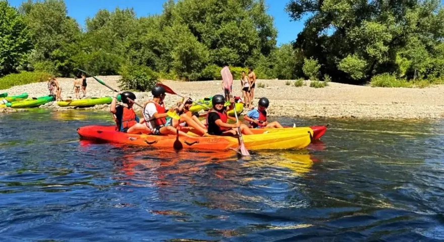 Des personnes profitent d'une activité canoe kayak sur une rivière ensoleillée, entourées de verdure et d'une plage de galets.
