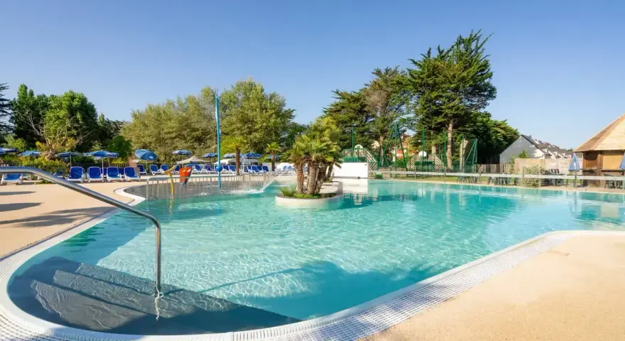 Piscine extérieure avec chaises longues bleues, parasols et végétation luxuriante sous un ciel bleu clair.