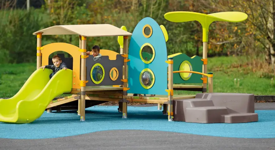 Des enfants jouent sur une aire de jeux colorée, respectant la réglementation aire de jeux extérieur. Un toboggan vert vif et une structure inspirée d'une fusée bleue.