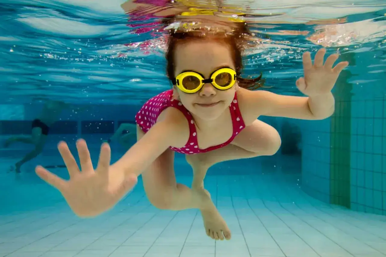 Une petite fille souriante, lunettes jaunes, nage sous l'eau. Elle est prête pour son attestation aisance aquatique.
