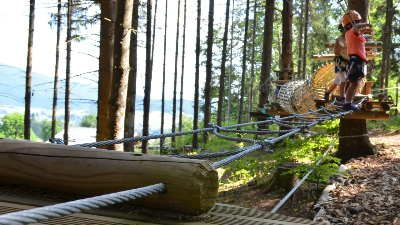 Des enfants s'amusent dans un parc aventure en Savoie. Ils traversent un pont de cordes, équipés de casques et de harnais, prêts pour leurs vacances scolaires.
