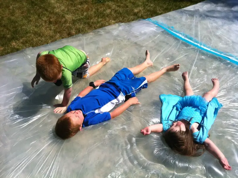 Trois enfants s'amusent sur un jeu d'eau pour kermesse, glissant sur une bâche mouillée sous le soleil.
