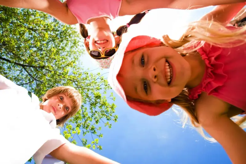 Trois enfants souriants, vus d'en bas, profitent d'une journée ensoleillée lors d'un camp sportif.